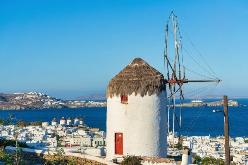 A white windmill with a red door, a thatched roof, and a large sail, overlooking a coastal town with white buildings and a clear blue sky. 1