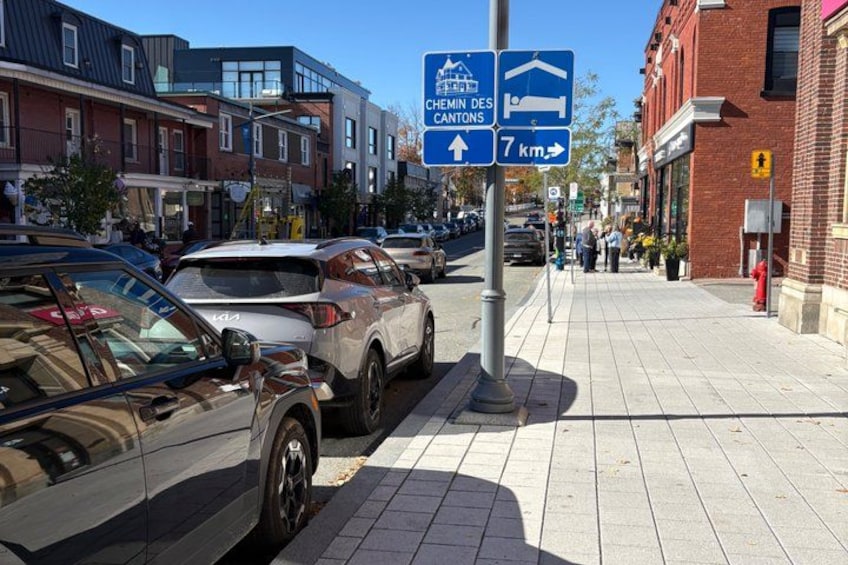 A street with parked cars, a pedestrian sidewalk, and a blue directional sign. 3