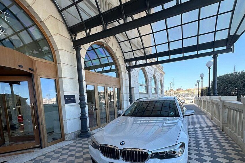 A white car parked in front of a building with a checkered floor pattern, large glass windows, and a covered entrance. 1