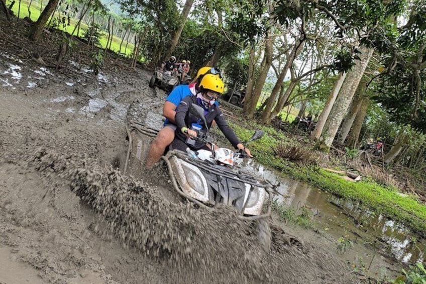 A person riding an ATV through a muddy area with trees and a small stream in the background. 2