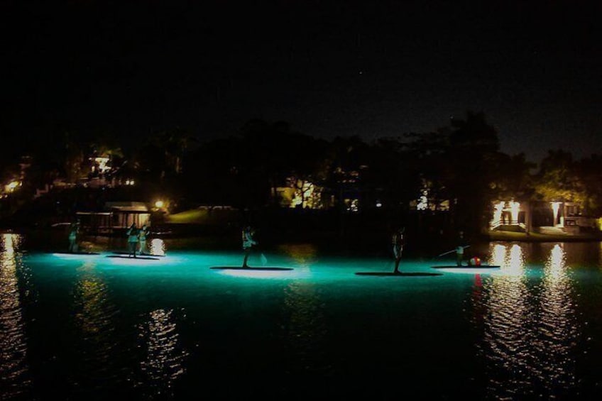 Ein nächtliches Bild mit ruhigem Gewässer, das von Umgebungslicht beleuchtet wird, und ein paar Personen auf schwimmenden Plattformen. 4