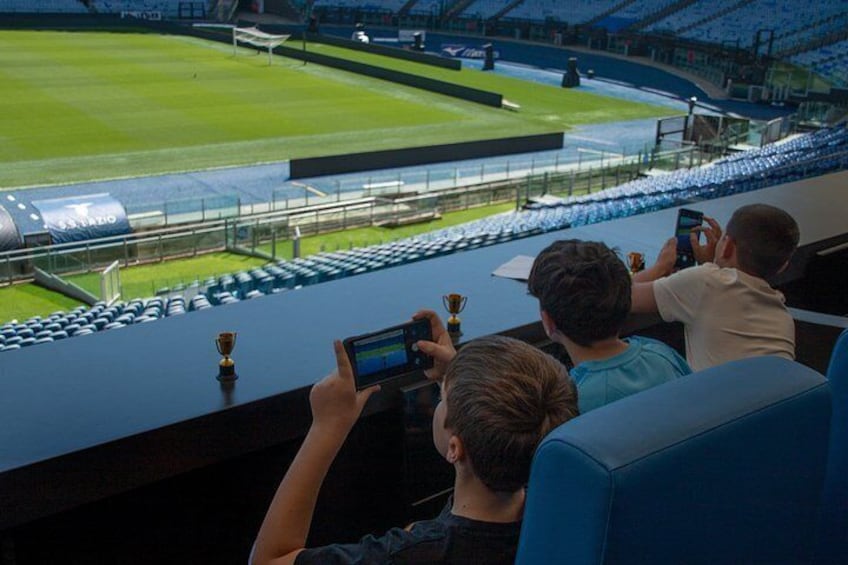 A stadium with empty seats, a green field, and a few people taking photos. 2