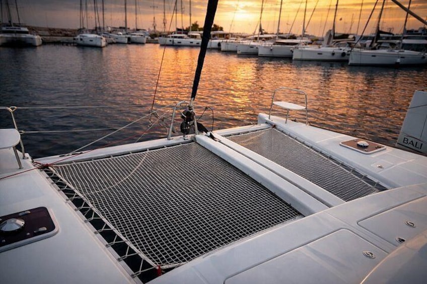 A sailboat with a white hull and a name 'BALI' on the side, docked at a marina with other boats and a sunset in the background. 1