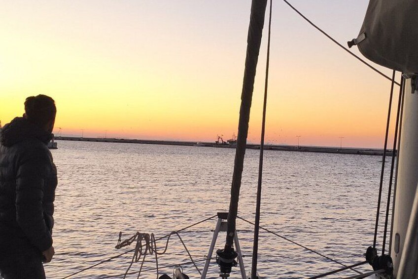 A person standing on a boat deck during sunset, with a calm sea and distant land visible. 2