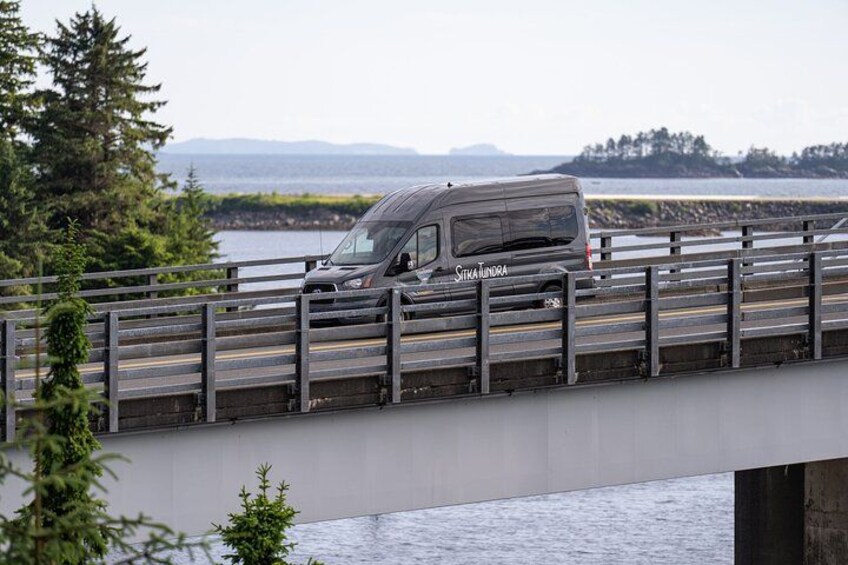 A van on a bridge with a body of water and trees in the background. 5