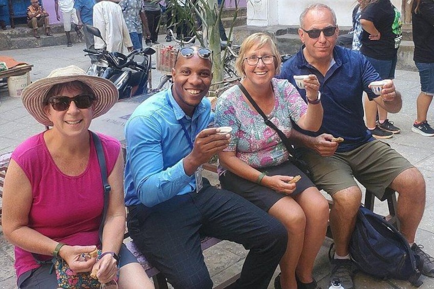 Cuatro personas sentadas en un banco, sosteniendo tazas, en un entorno al aire libre con otras personas y bicicletas de fondo. 2