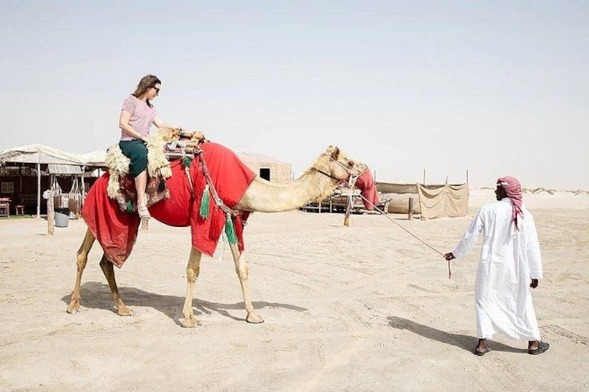 Eine Person reitet auf einem Kamel durch eine Wüste, im Hintergrund sind Zelte zu sehen. 2