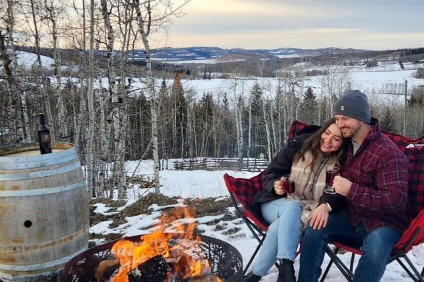 Una pareja sentada junto a una fogata en un paisaje nevado con árboles y una vista distante. 4