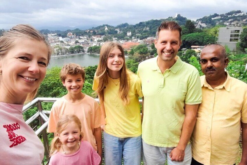 Eine Gruppe von Menschen steht auf einem Balkon mit Blick auf eine Stadtlandschaft, im Hintergrund Fluss und Hügel. 3