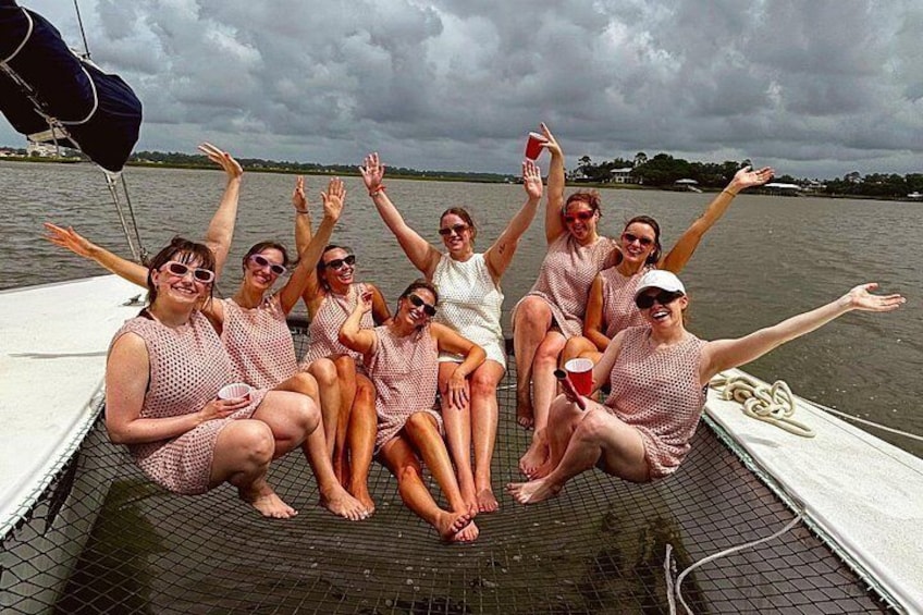 A group of people on a boat, some wearing pink dresses, with a body of water and a cloudy sky in the background. 2