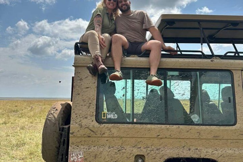 Deux personnes sont assises sur le toit d’un véhicule de safari, avec un paysage verdoyant en arrière-plan. 3
