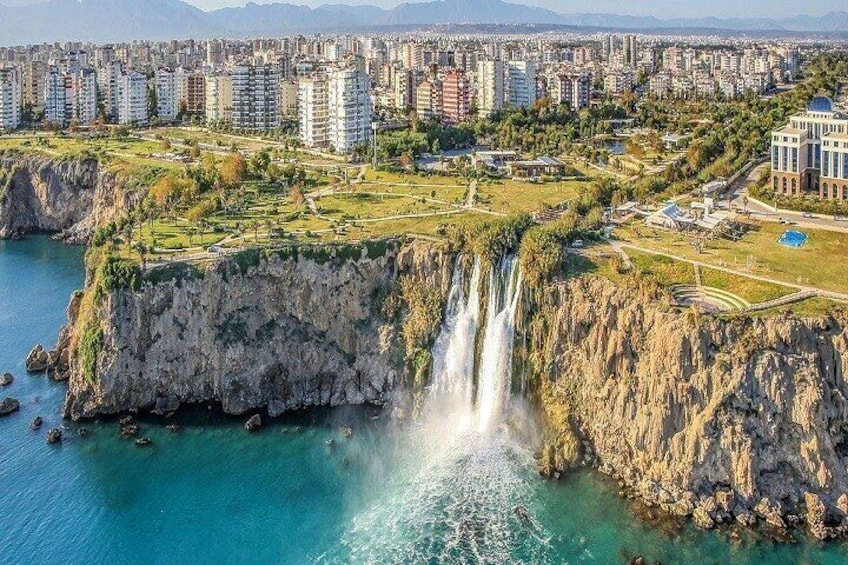 A coastal cityscape with a prominent waterfall cascading into turquoise waters, surrounded by residential buildings and greenery. 1