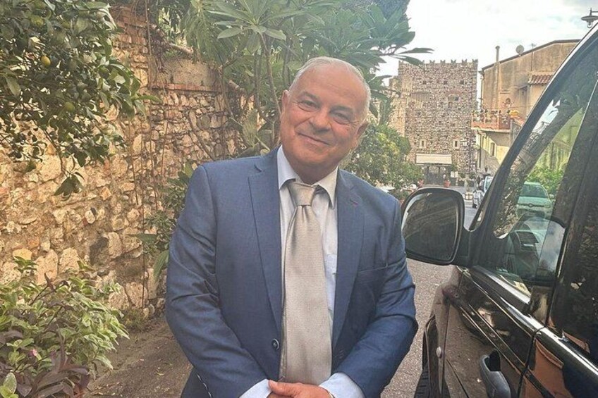 A man in a blue suit standing next to a black car in a narrow street with stone walls and greenery. 2
