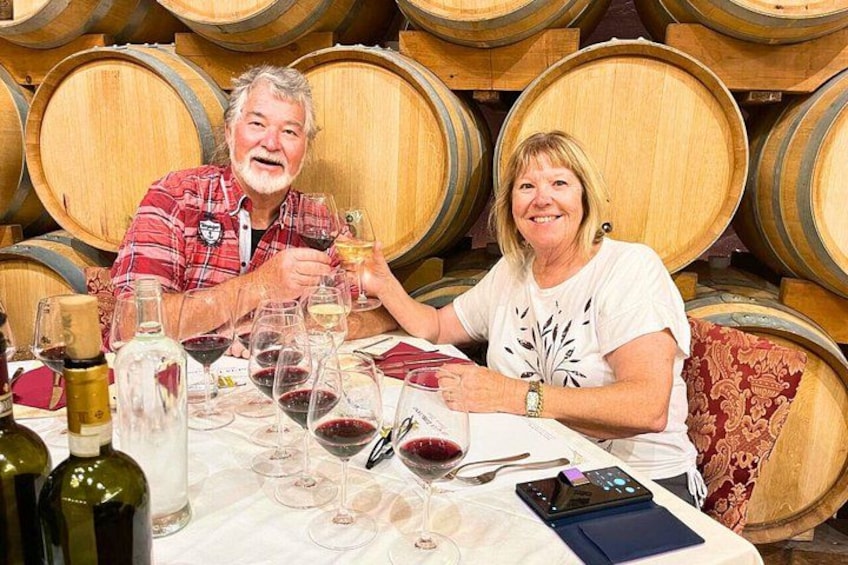 Two people seated at a table with wine glasses and bottles, surrounded by wooden barrels. 2
