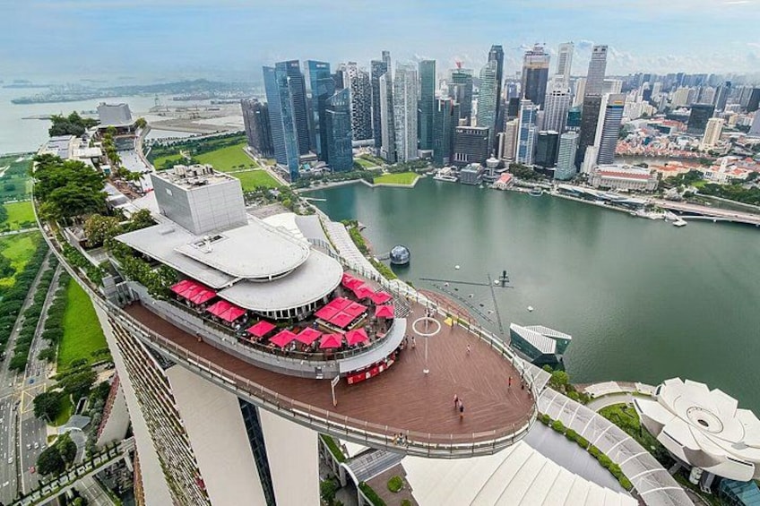 A waterfront promenade with a circular viewing platform, a modern building with a white facade, and a city skyline in the background. 3