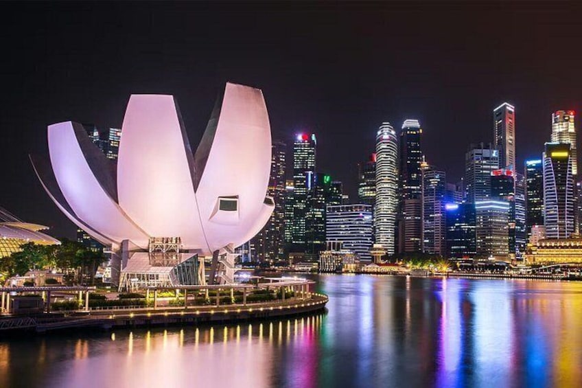 A city skyline at night with illuminated buildings and a lotus-shaped structure reflecting on the water. 1