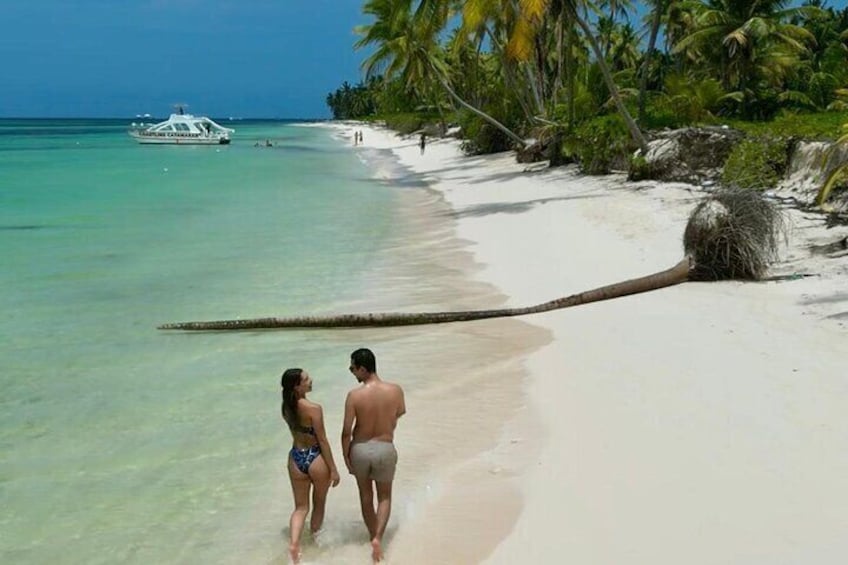 Una coppia che cammina su una spiaggia con acque turchesi cristalline, una barca all'orizzonte e palme. 2