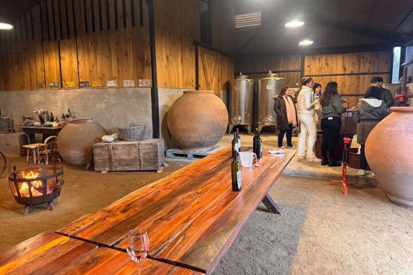 A wooden table with bottles and a wine glass, surrounded by large ceramic pots and barrels in a rustic setting. 5
