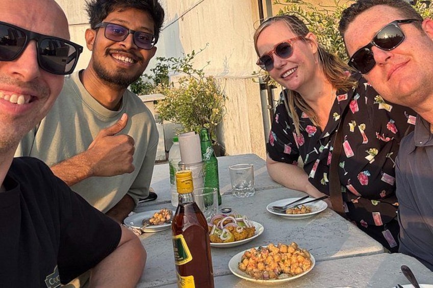 Vier personen zitten aan een buitentafel met borden eten, een fles saus en een glas water. 3