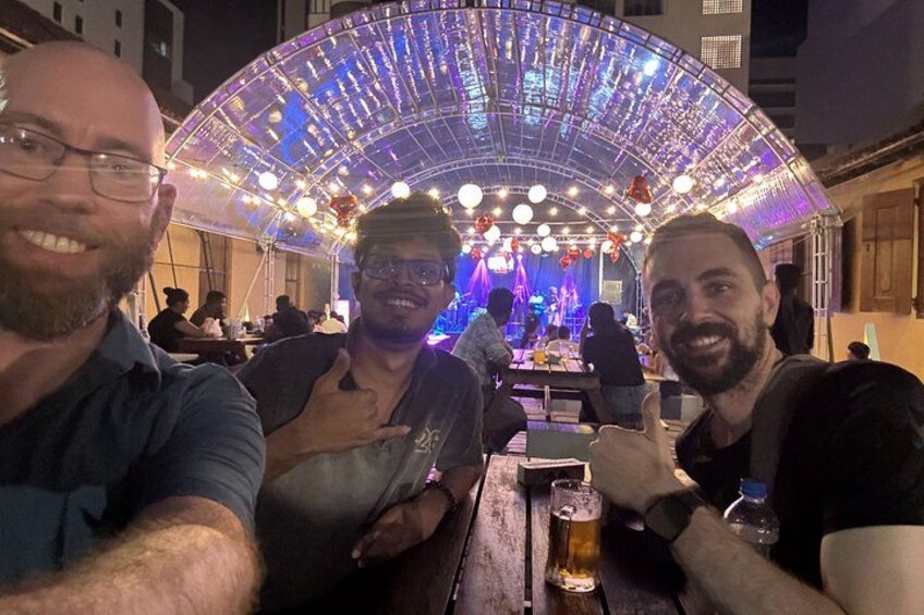 Drie personen zitten aan een tafel met drankjes, onder een luifel met lichtjes. 2
