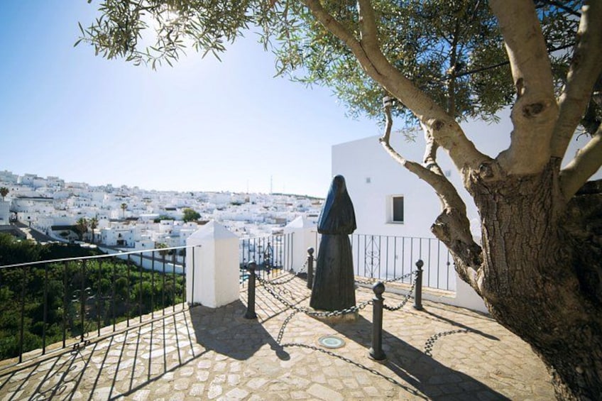 A statue on a stone-paved area with a view of a white building and a tree. 2
