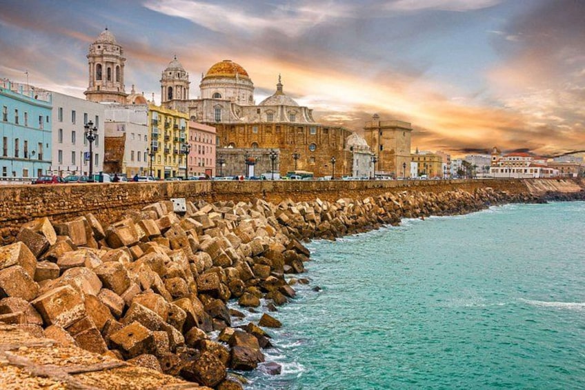 A coastal cityscape with a prominent historical building with domes, a rocky breakwater, and a clear sky. 1