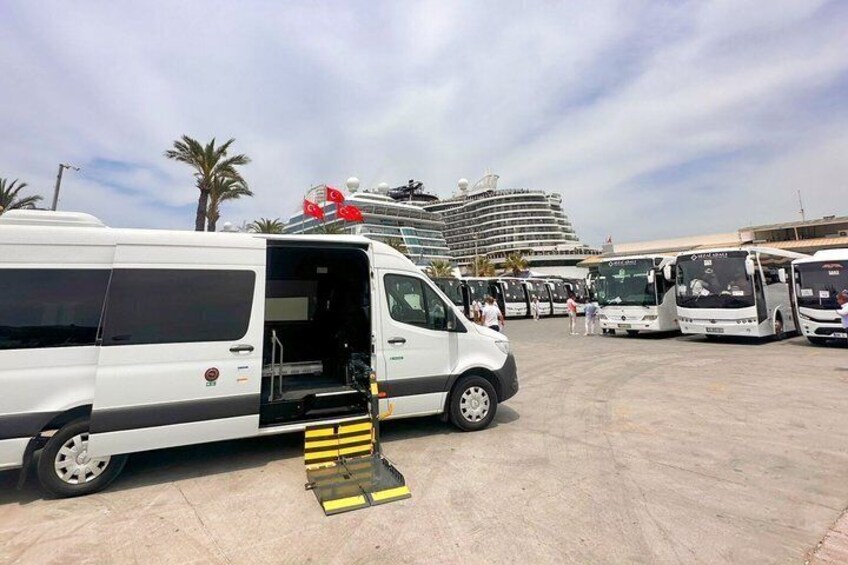 Een witte bestelwagen met geopende deur, een hellingbaan en meerdere bussen geparkeerd op een parkeerplaats, met een cruiseschip op de achtergrond. 2