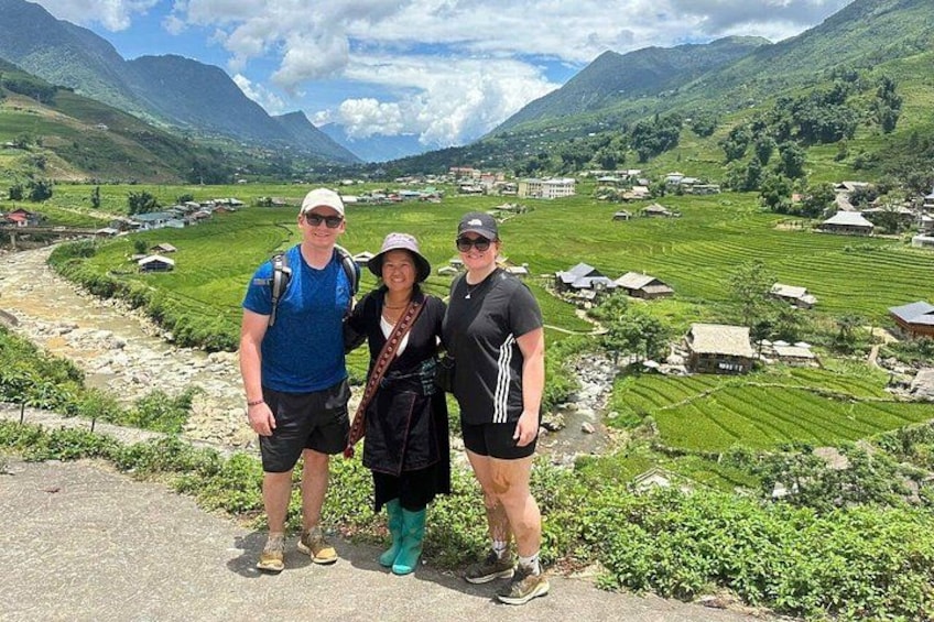 Drie personen staan op een pad, met een bergachtig landschap en terrassen in de achtergrond. 2