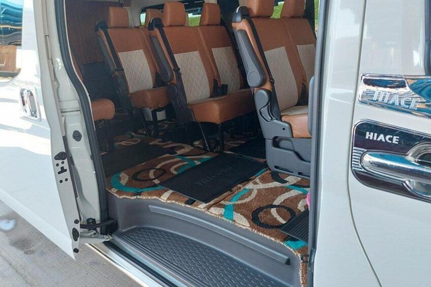 Interior de una camioneta con asientos marrones y color beige, un felpudo estampado y el logotipo 'HIACE' en la puerta. 4