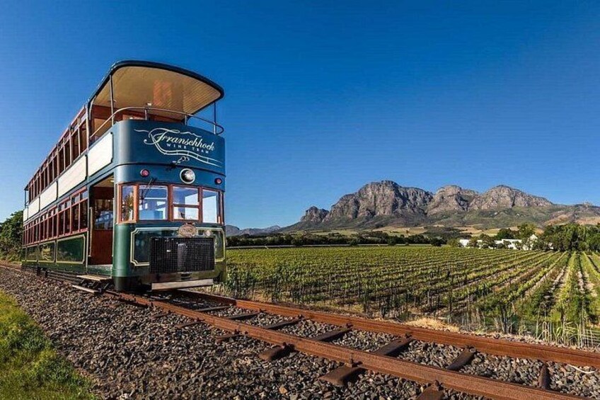 Een klassieke tram met de tekst 'Franschhoek' er op, rijdt over rails met wijngaarden en bergen op de achtergrond. 5