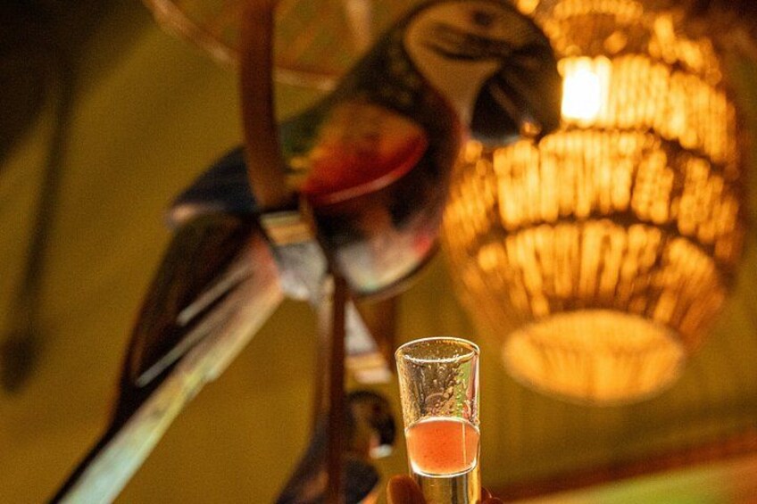 Een cocktailglas met een roze vloeistof, een beeld van een vogel, en een warme, onscherpe achtergrond met een kroonluchter. 5