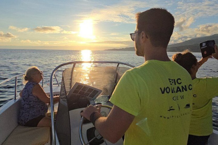 A boat with people on board, a sunset in the background, and a person taking a photo. 1
