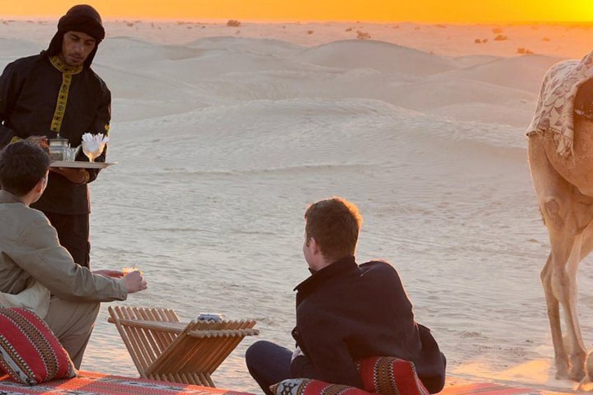 A desert landscape with a camel, a person serving food, and two people sitting on a blanket. 1