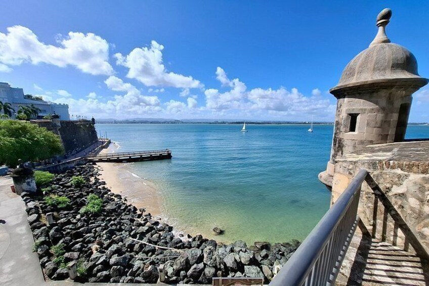 Blick auf die Küste mit einem Steinturm, klarem, blauem Wasser, einer felsigen Uferlinie und einem entfernten Segelboot. 1