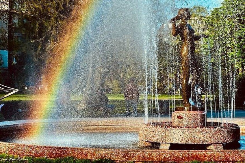 A fountain with water jets and a statue, surrounded by trees and a rainbow. 5