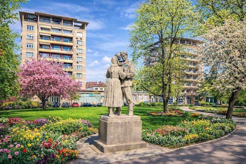 A statue of a couple in a park with flowering trees and a building in the background. 1