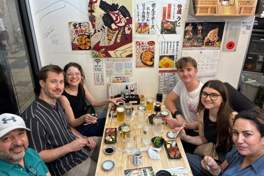 A group of people sitting around a table with food and drinks, in a room with various posters and signs on the walls. 5