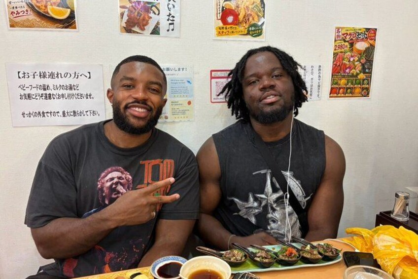 Two people sitting at a table with food, a menu, and a wall with various posters. 2