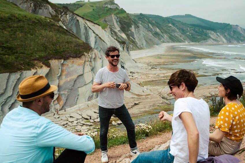 A group of people sitting and standing on a rocky cliff overlooking a beach with cliffs in the background. 1
