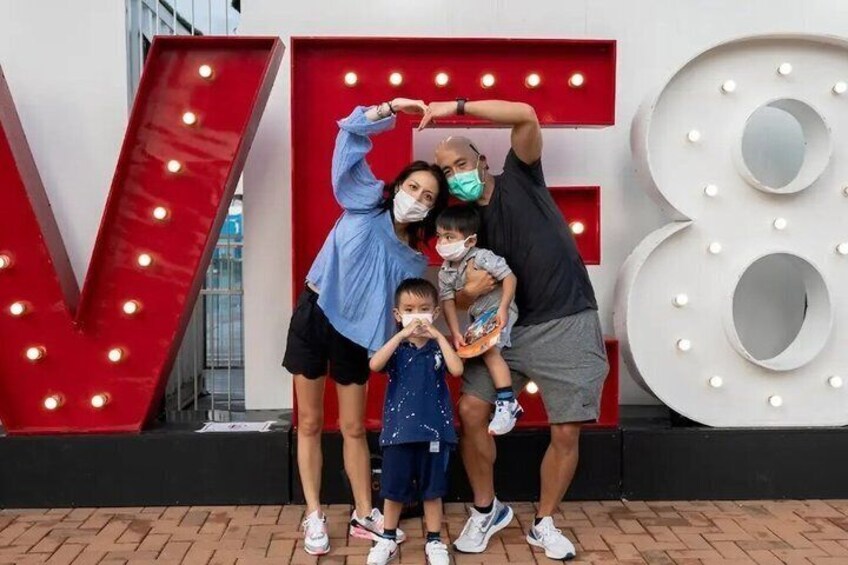 A family posing in front of large illuminated letters 'V' and '8' with a brick pavement. 1