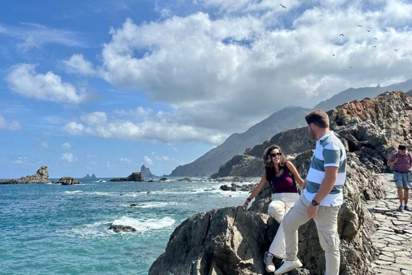 A rocky coastline with clear blue water, a person standing on a rock, and another person sitting on a rock. 2