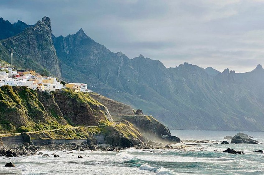 A coastal village on a cliff, rugged mountains in the background, and waves crashing against the shore. 1