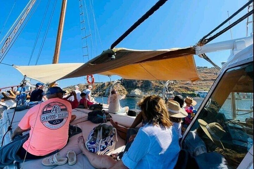 A group of people on a sailboat with a clear blue sky and rocky coastline in the background. 2