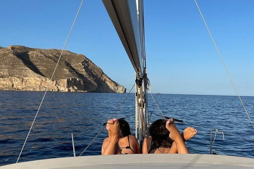 A sailboat with two people reclining on the deck, a rocky coastline, and calm blue waters. 5