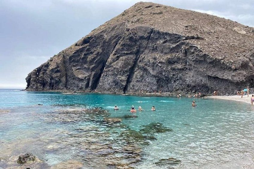 Une côte rocheuse bordée d’une eau turquoise cristalline, où l’on voit des gens se baigner, et une grande colline en arrière-plan. 2