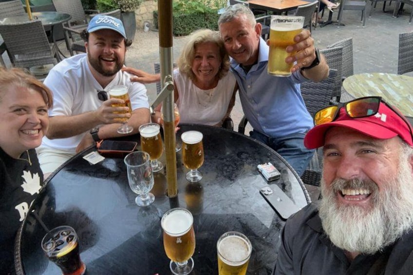 Un grupo de personas sentadas alrededor de una mesa redonda, con vasos de cerveza, una parrilla y mobiliario de exterior. 2