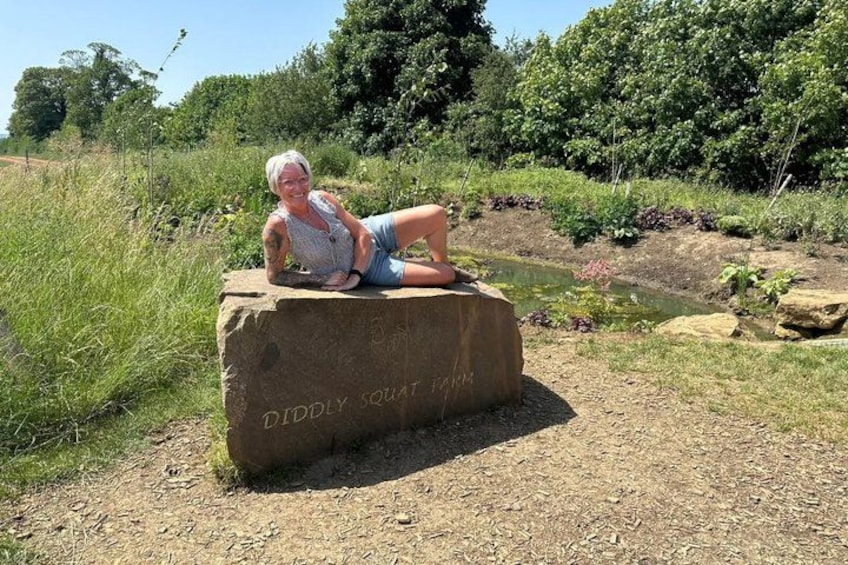 Una persona sentada sobre una roca con la leyenda 'DIDDLY SQUAT FARM' en un entorno natural con césped y árboles. 3