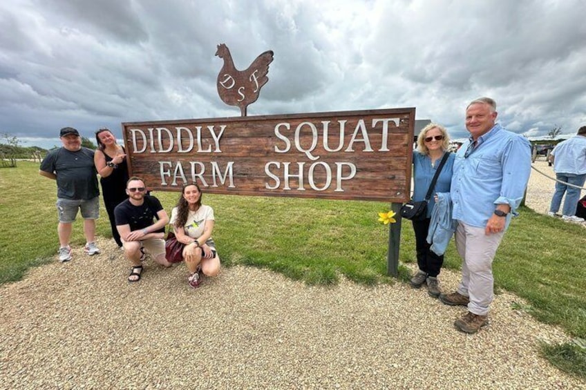 Un grupo de personas posando frente a un letrero de madera que dice 'DIDDLY SQUAT FARM SHOP' con un logotipo de gallina. 1