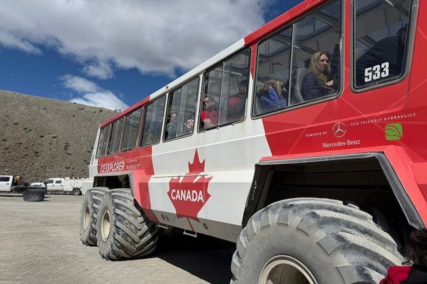 A red and white tour bus with 'CANADA' and 'MERCEDES-BENZ' branding, parked on a gravel surface with a mountainous background. 4