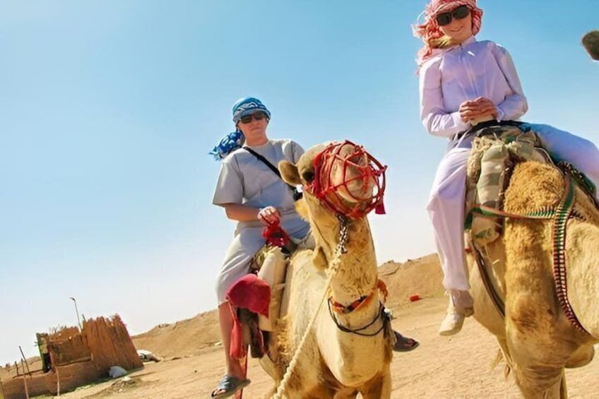 Two people riding camels in a desert landscape with sand dunes and a clear sky. 1